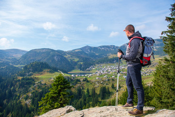 Hiker young man with backpack and trekking poles standing on edge of cliff and looking at the lake,...