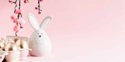 Easter golden eggs, pink flowers and porcelain rabbit on pastel pink background. Easter, spring concept. Copy space, banner