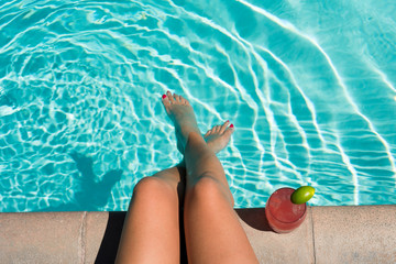 Woman dips her legs in a pool and sips on a cocktail drink