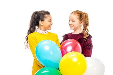 Fototapeta na wymiar smiling schoolgirls holding colorful balloons and looking at each other isolated on white