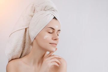 Skin care. A cute young  girl with a towel on her head puts a nourishing cream on her face, her...