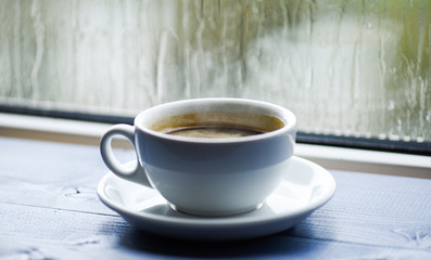 Wet glass window and cup of hot coffee. Autumn cloudy weather better with caffeine drink. Enjoying coffee on rainy day. Fresh brewed coffee in white cup or mug on windowsill. Coffee time on rainy day