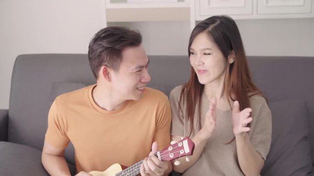 Asian couple playing guitar and singing together in living room at home, sweet couple enjoy love moment while lying on the sofa when relax at home. Lifestyle couple relax at home concept.