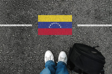  a man with a shoes and backpack is standing on asphalt next to flag of Venezuela and border