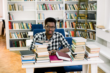 Ethnic african american guy sitting at table surrounded by books in library. Student is talking on phone