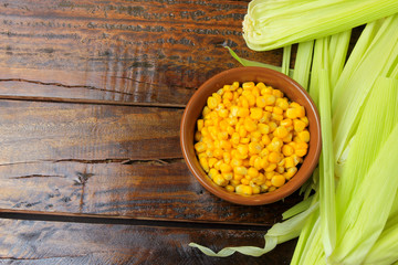 Raw corn kernels, inside ceramic bowl, next to corn on the cob on rustic wooden table.