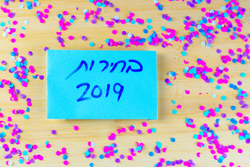 Hebrew text Elections 2019 on voting paper over wooden board with confetti background. Israeli legislative Elections for the 21st Knesset Israel 9 April 2019.