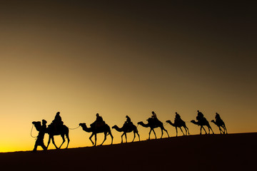 Sillhouette of camel caravan with happy peopple going through the desert at sunset with red sky in background