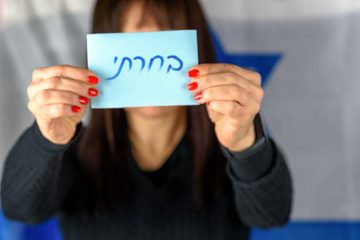 Young Woman Holding Ballot Front Of Face on Israeli Flag Background.Hebrew text I Voted on voting paper.