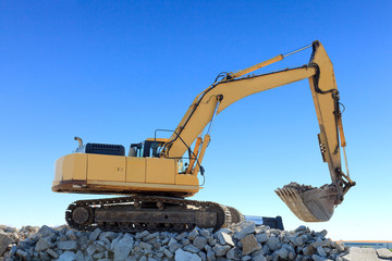 Construction backhoe on top of a mound of rocks. Bright blue sky is in the background.