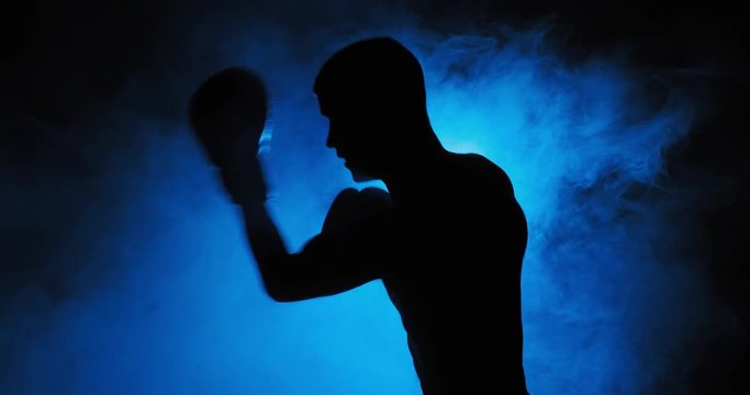 Muscular young man in boxing gloves shows the different movements and strikes in the studio on a dark background. Fog in the background