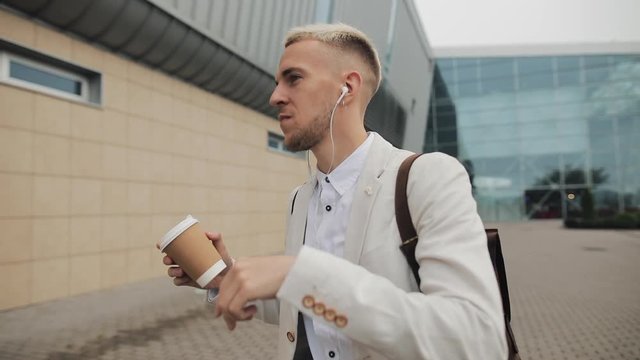 Happy young businessman with coffee listening to music in headphones on smartphone and funny dancing walking near office building. Funny dancing moves. Slow motion
