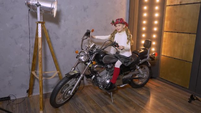 Studio portrait of pretty young girl in hat sitting on motorcycle in stodio and photographing at lights background