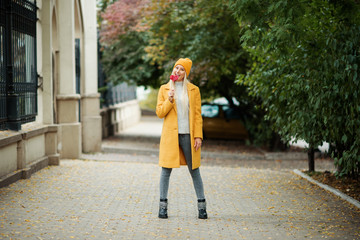 Saint Valentine's day concept. Fashion portrait blond young woman in yellow coat having fun with red lollipop heart over street background. 