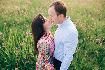 Beautiful young couple gently hugging and kissing in sunshine in fresh spring meadow with pink flowers. Happy stylish family embracing in green field. Romantic moments. Valentine day