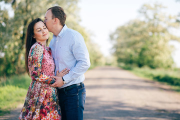 Beautiful young couple sensual hugging and kissing on road in sunshine among spring field and trees. Happy family in love embracing in sunlight. Romantic joyful moments. Space for text