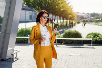 Attractive business woman in sunglasses and yellow suit walking near office centre with cup of coffee and uses smart phone. Business lifestyle and success concept. Lunch break among working day.