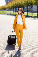 Beautiful business woman in stylish yellow suit pulls a suitcase, hurries to the airport and talks on smart phone. Attractive young woman going on a business trip pulling her suitcase behind her.