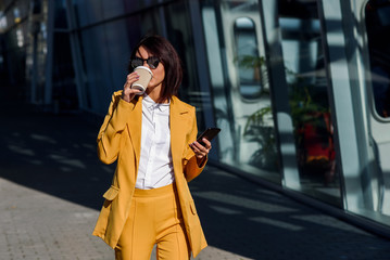 Beautiful business woman in yellow suit and sunglasses walking near business centre, drinks coffee and uses smartphone. Business concept, communication, manager and office worker.