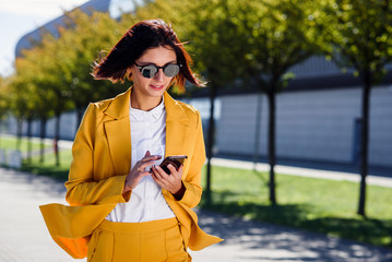 Stylish young business woman uses smart phone while walks near business centre on alley background....
