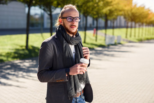 Young Caucasian Businessman With Backpack And Cup Of Coffee Outdoors Near Modern Business Center. Young Bearded Businessman Walking On The Street Drinks Coffee During Lunch Break.