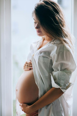 Young pregnant woman standing near window at home and thinking about the future. Smiling pregnant...