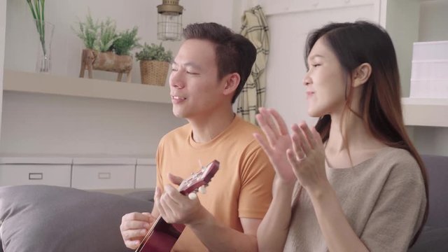 Asian couple playing guitar and singing together in living room at home, sweet couple enjoy love moment while lying on the sofa when relax at home. Lifestyle couple relax at home concept.