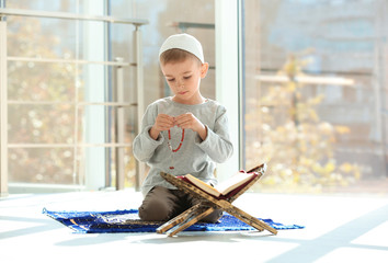 Little Muslim boy with misbaha and Koran praying on rug indoors