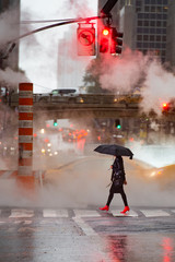 A woman with an umbrella and red high heels shoes is crossing the 42nd street in Manhattan. Taxi and steam coming out from from the manholes in the background. New York City, Usa.