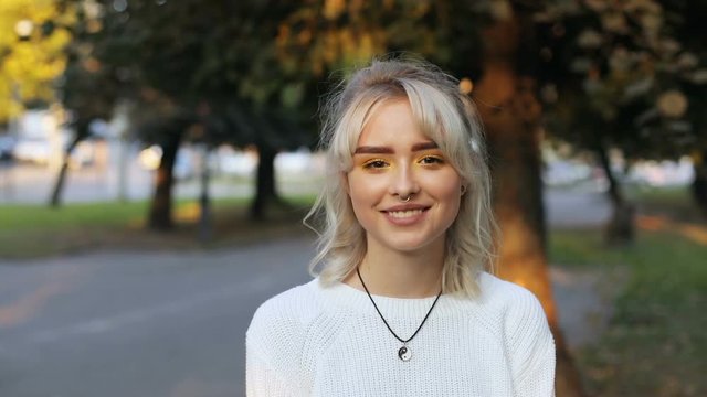 Portrait of good-looking young woman with blonde hair looking at camera and smiling. Cute girl with septum piercing. Green park in background.