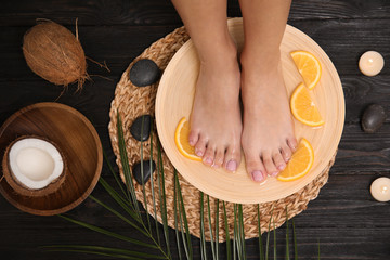 Woman soaking her feet in plate with water and orange slices on wooden floor, top view. Spa...