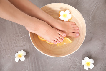 Obraz na płótnie Canvas Woman soaking her feet in bowl with water, orange slices and flower on grey background, top view. Spa treatment
