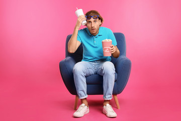 Emotional man with 3D glasses, popcorn and beverage sitting in armchair during cinema show on color...
