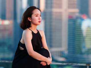 Fototapeta na wymiar Beautiful young brunette woman in sexy black dress sitting on top of mansion roof with blur Shanghai Bund landmark buildings background with dusk light. Emotions, people, beauty and lifestyle concept.