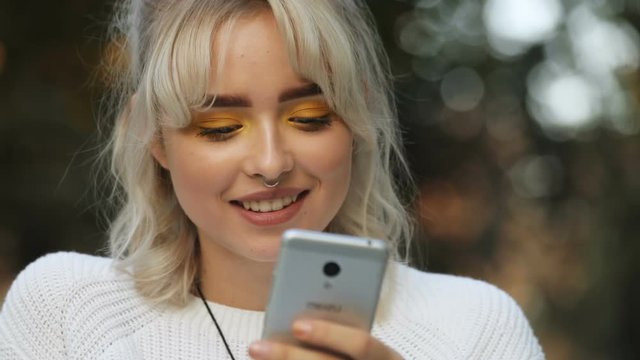 Pretty Caucasian young woman using smartphone, smiling. Portrait of cheerful girl with septum piercing and yellow eye shadow. Close-up. Blonde hair. Daytime.