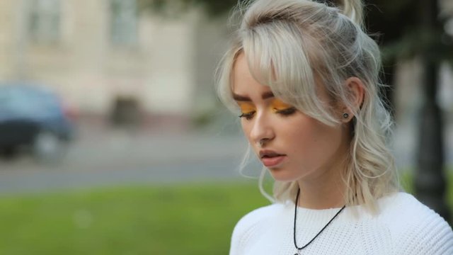 Close-up portrait of cute hipster in street. Young focused woman with septum piercing and yellow eye shadow looking down. Outdoors.