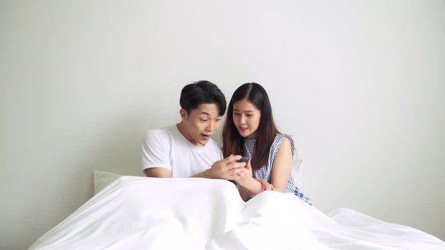 Couple in bed looking at mobile phone with winning posture. Asian chinese man and woman in bed in day time with laptop. Modern lifestyle concept.