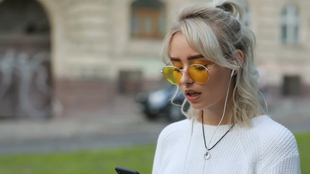 Awesome girl with blonde hair holding black smartphone in hand, wearing headphones and listening to music. New songs, playlist. Outdoors.