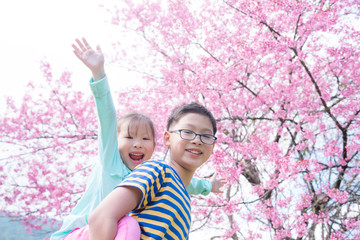 Little happy asian girl with her brother playing in front of blooming pink flower cherry tree.