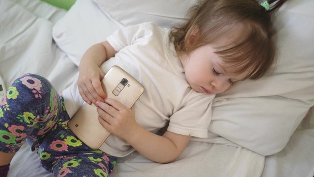 child is sleeping on pillow and holding tablet. Cute baby sleeping in bed with smartphone.