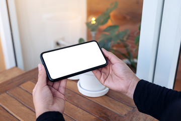 Mockup image of hands holding and using a black mobile phone with blank screen horizontally for watching in the outdoors