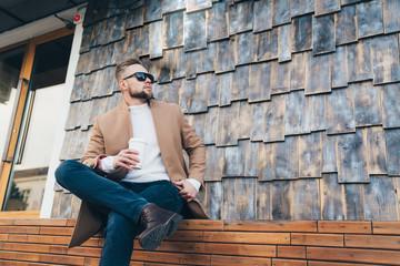 Handsome man in stylish clothes sitting near cafe with his legs crossed