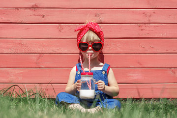 Little girl in dungarees and sun glasses is drinking milk. Colorful summer concept.