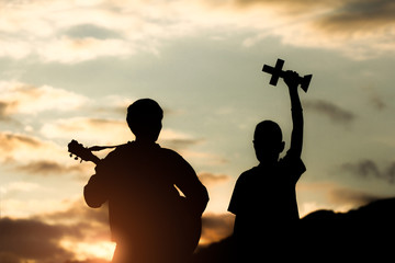 Two young christian worship God by holding cross and playing guitars with light sunset...