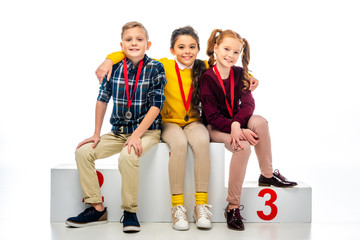 smiling kids with medals sitting on winner podium and looking at camera isolated on white