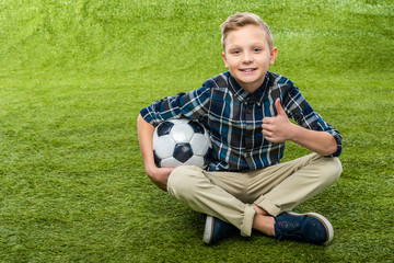 smiling boy holding soccer ball, looking at camera and showing thumb up on lawn