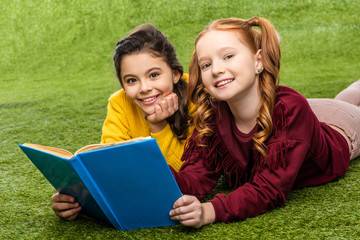 smiling schoolgirls lying on lawn, holding book and looking at camera