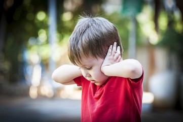 A 4 year old autistic child in a red shirt closing his ears with hands as if protecting from noise....