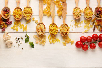 Spoons with different types of raw pasta on white wooden background
