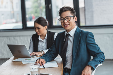 young kazakh businessman in eyeglasses smiling at camera while female colleague using laptop behind in office